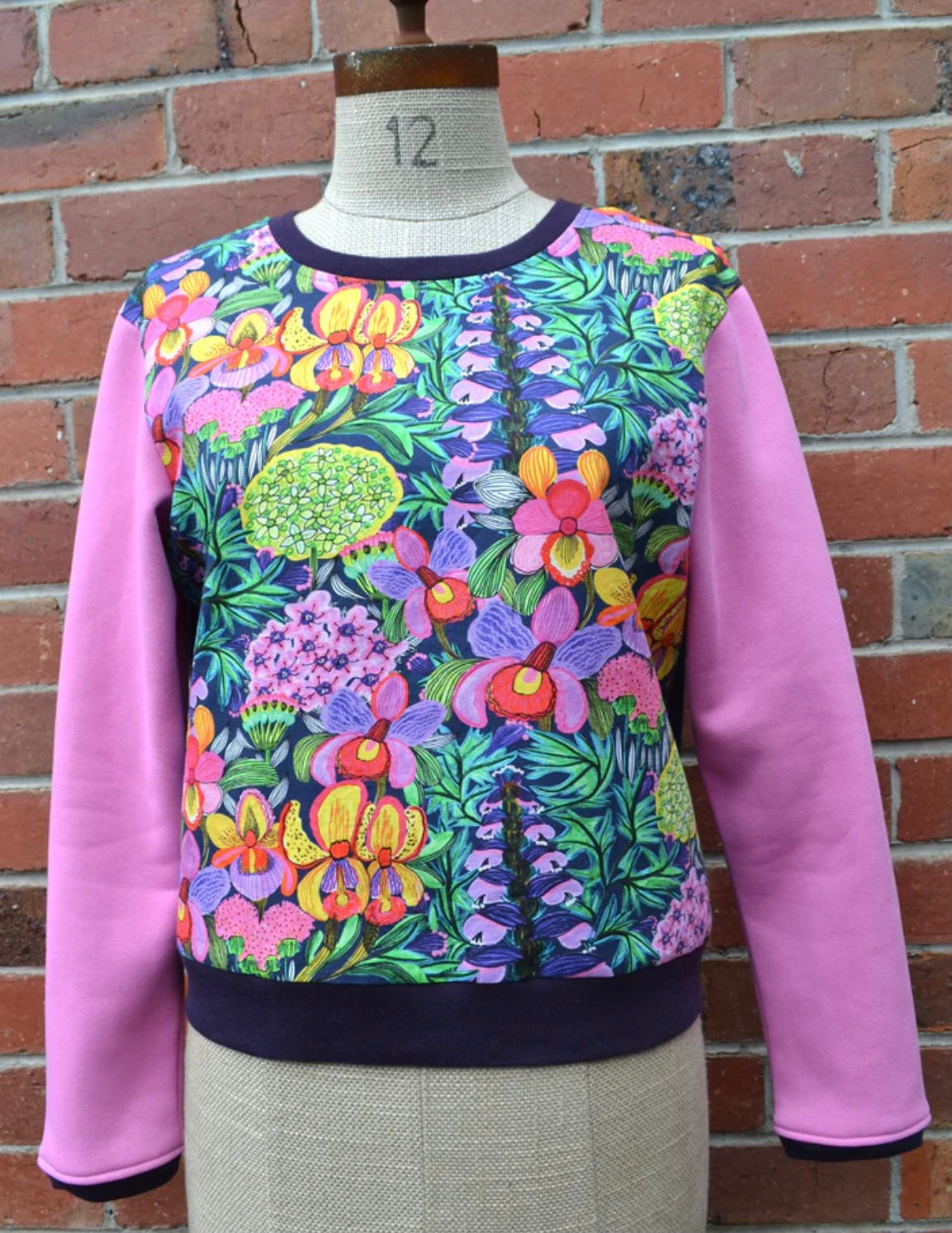 Colorful floral-patterned sweater with pink sleeves on a mannequin against a brick wall.