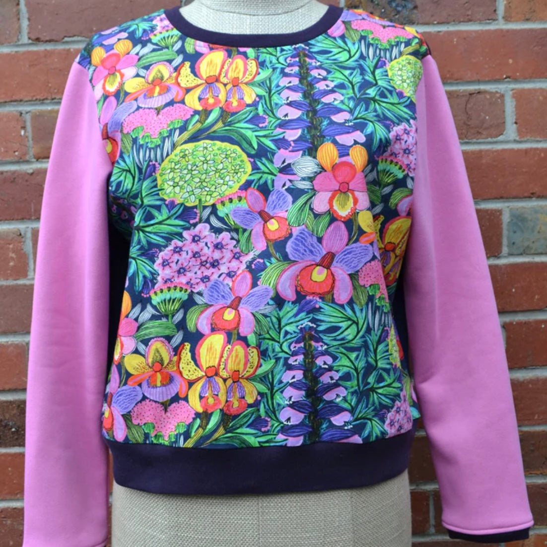 Colorful floral-patterned sweatshirt with pink sleeves against a brick wall.