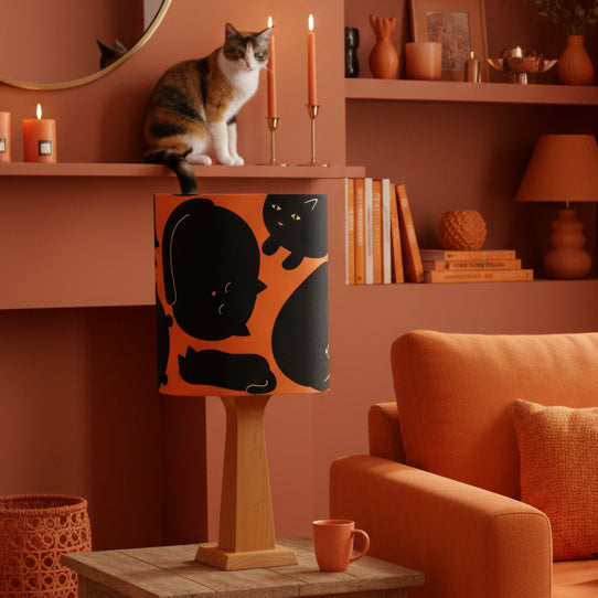 A table lamp with a cylindrical shade featuring a black cat pattern on an orange background, supported by a light brown wooden pedestal base.