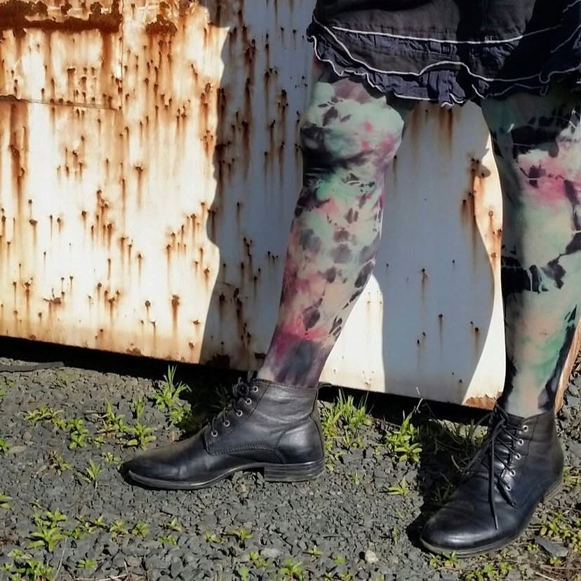 Person wearing black shoes and colorful tie-dye leggings standing near a rusty metal surface.