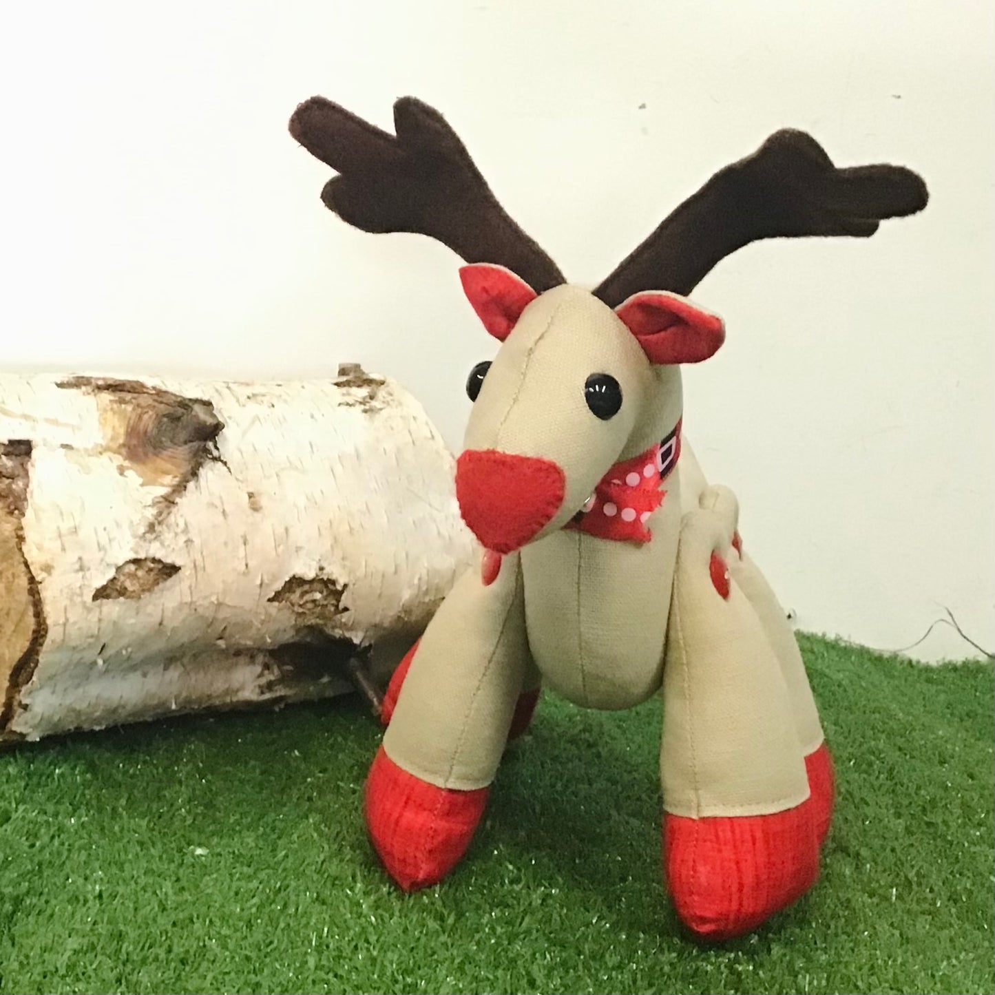 Plush reindeer toy with red nose and antlers on a grassy surface with a log in the background