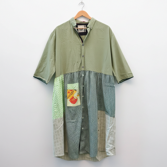 A patchwork short-sleeve button-up shirt hanging on a wooden hanger against a white brick wall. The shirt features multiple fabric panels in sage green, blue-green gingham, olive green, and a small colorful patch on the front pocket.