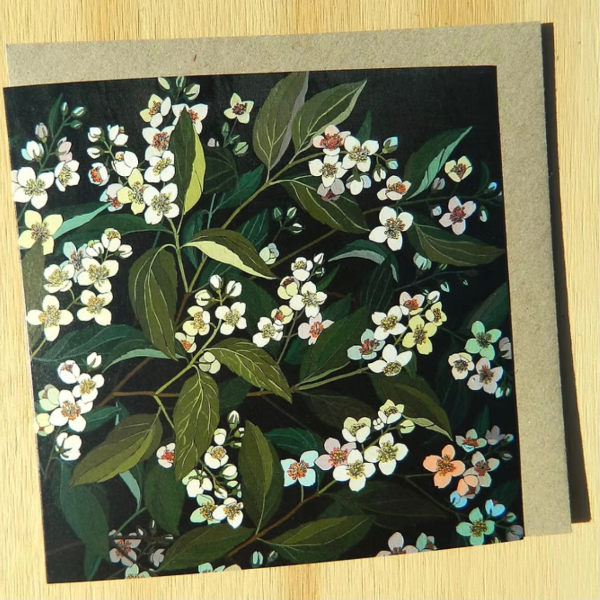 Floral card with white flowers and green leaves on a dark background, placed on a wooden surface.