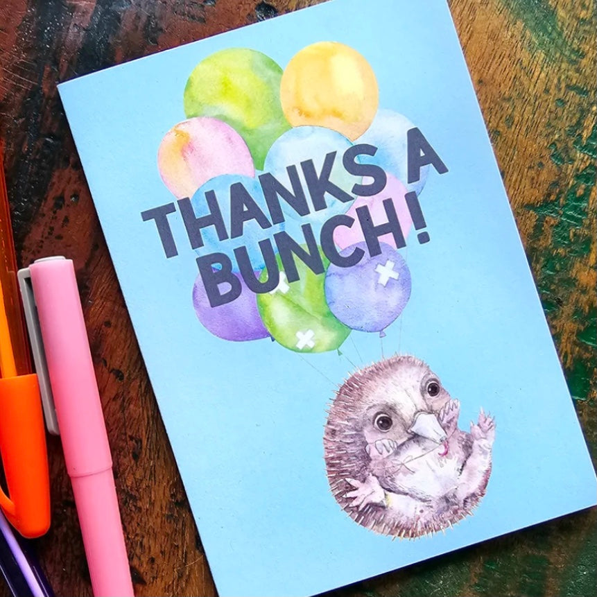 greeting card with 'Thanks a Bunch!' text and balloons on a wooden surface with pens.