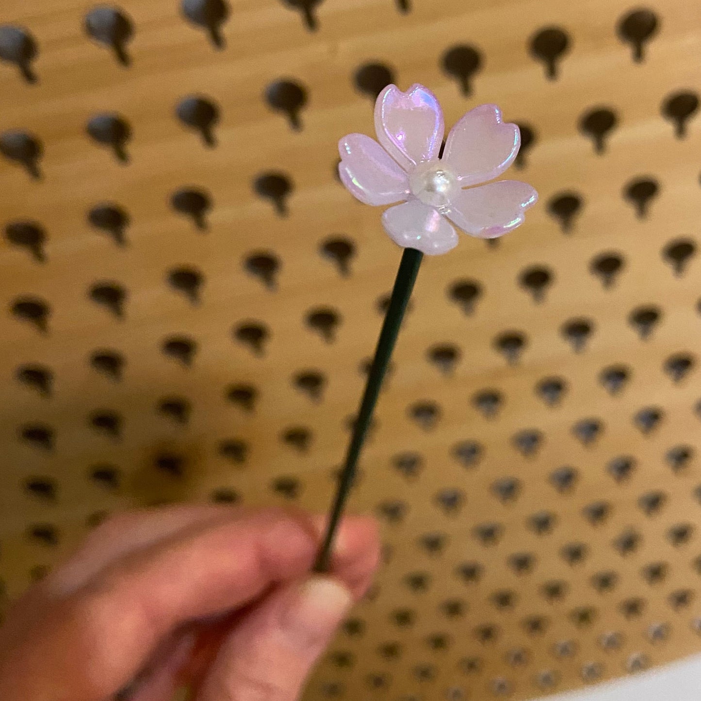 Hand holding a single flower stem with a purple flower design against a wooden pegboard background