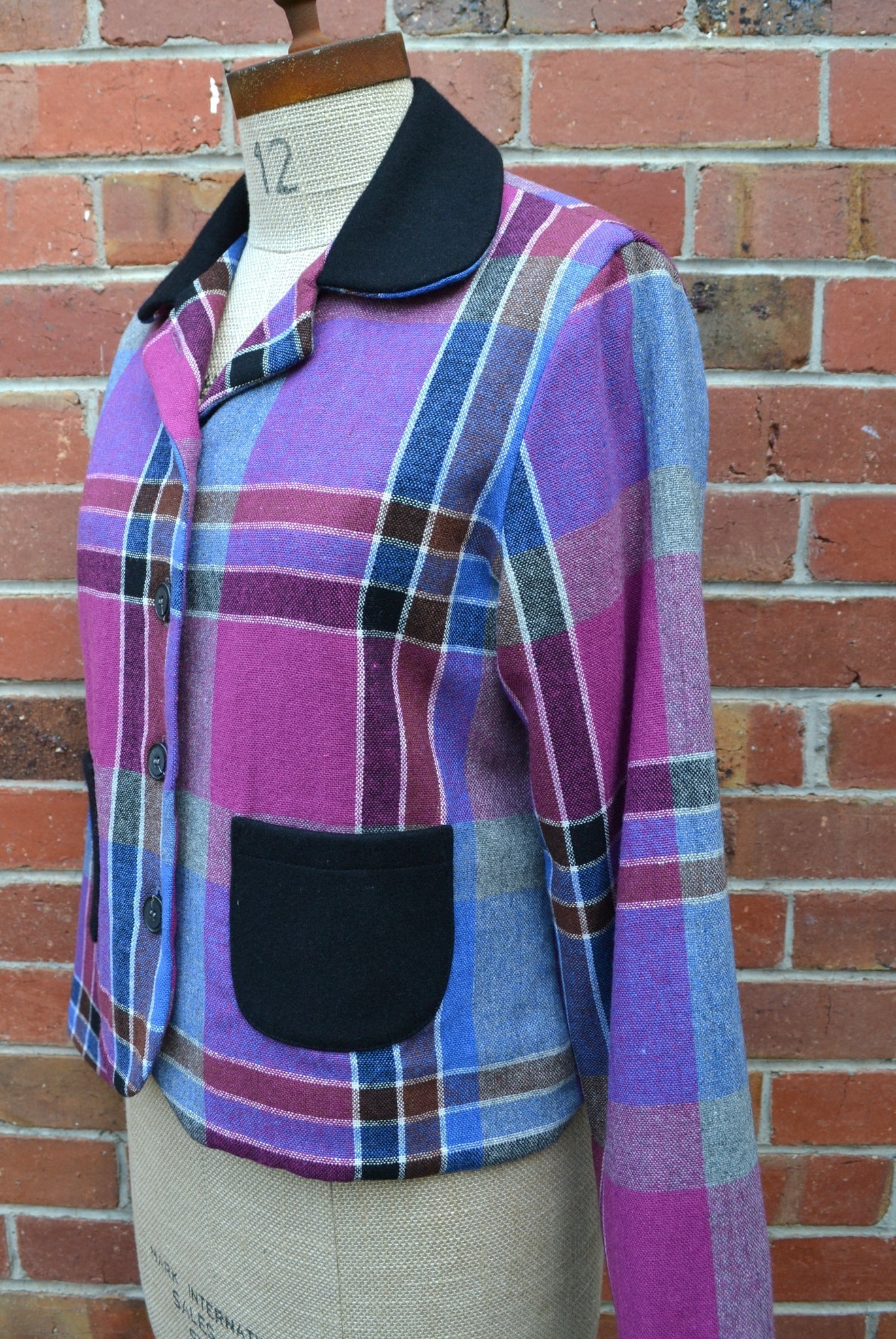 Plaid cropped jacket with a black pocket on a mannequin against a brick wall.