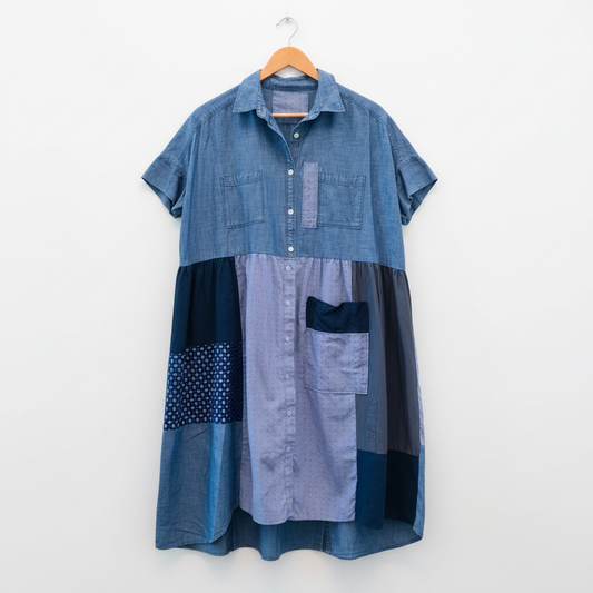 A patchwork denim shirt dress hanging on a wooden hanger against a plain white wall. The dress features a medium blue denim upper section, a light blue patterned middle section, and dark navy blue patchwork panels with polka dots and solid fabric.