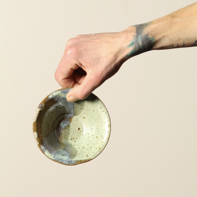 Hand holding a ceramic bowl with a speckled glaze on a beige background