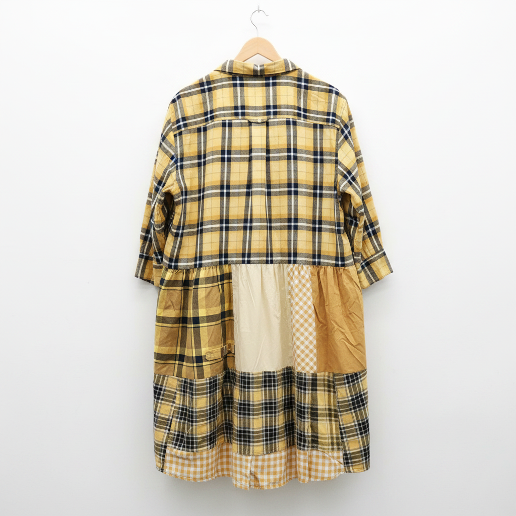 A patchwork plaid shirt dress hanging on a wooden hanger against a white-painted brick wall. The dress features a cream, black, and tan plaid pattern with solid tan and checkered fabric panels.