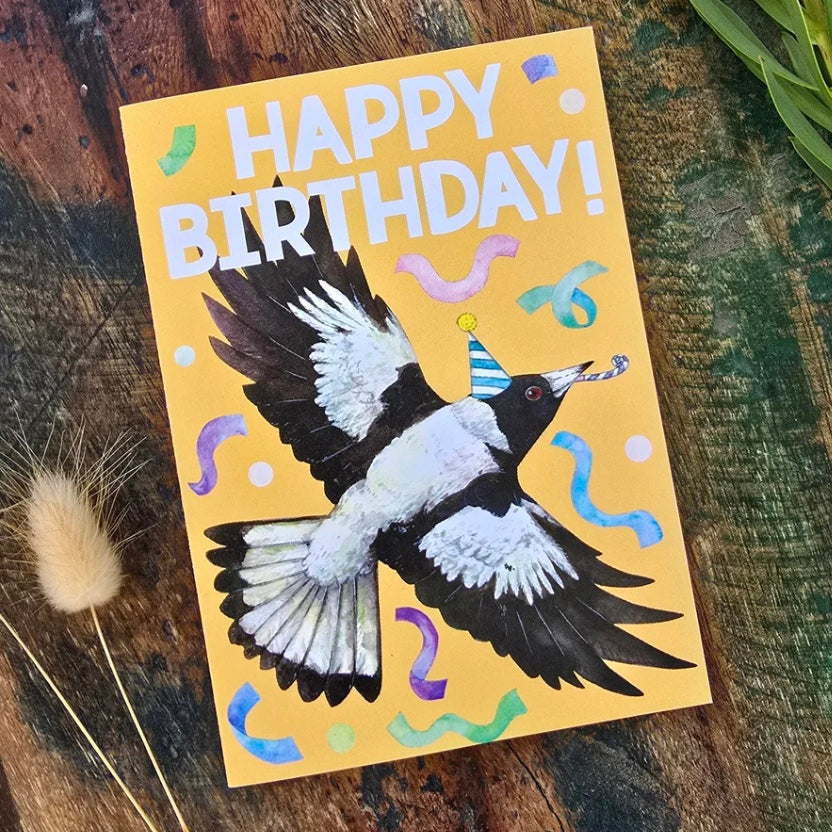 Birthday card with a MAGPIE bird illustration and 'Happy Birthday!' text on a wooden surface.