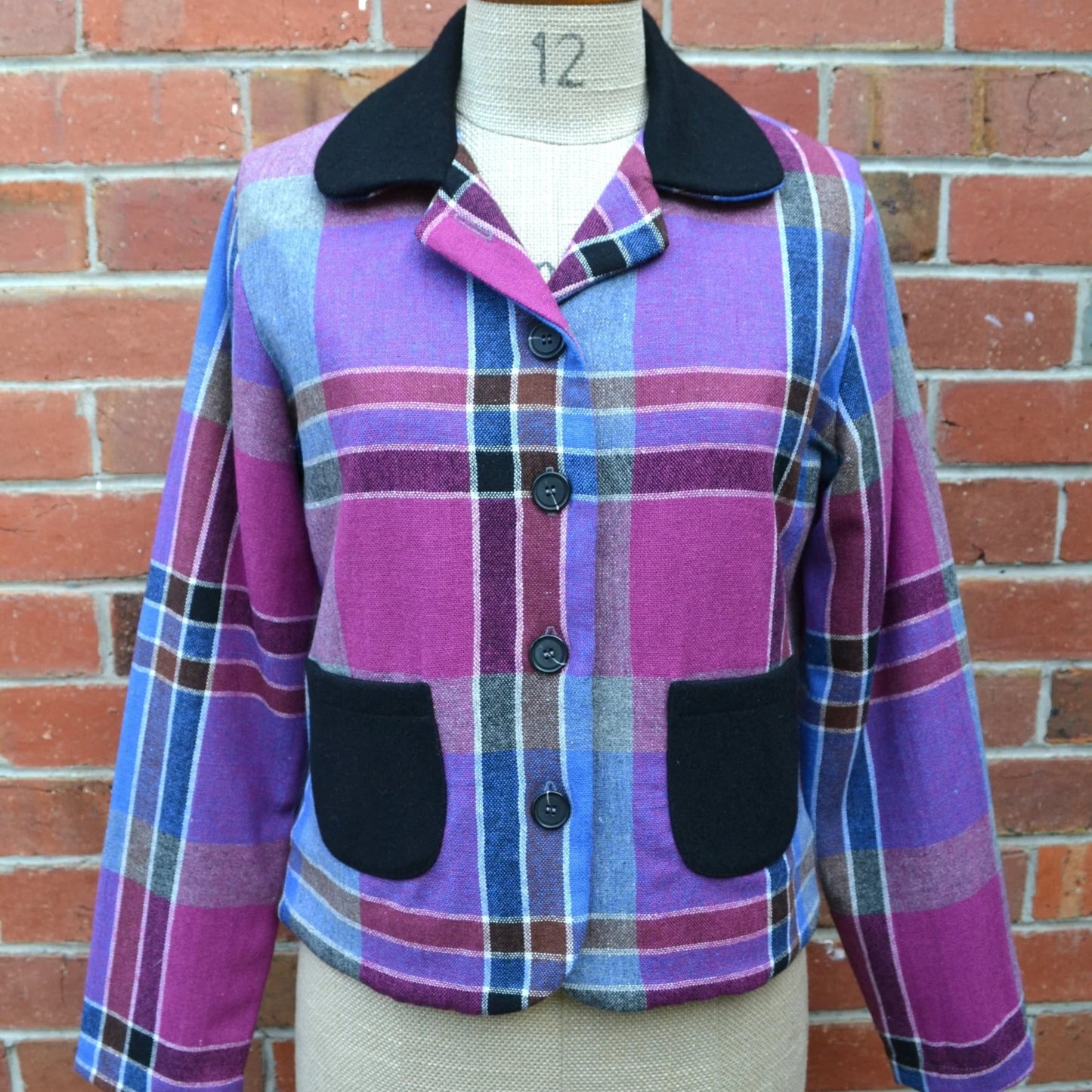 Plaid cropped jacket with purple and pink pattern and black collar and pockets on a mannequin against a brick wall.