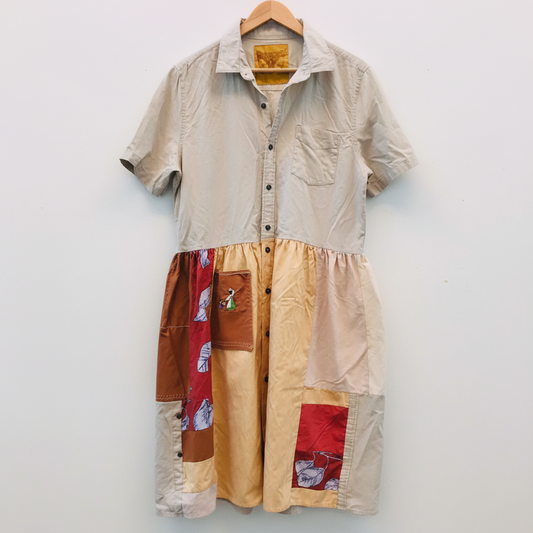 A short-sleeve button-front shirt dress in a light beige linen-like fabric, featuring a patchwork lower half with brown, red, and cream fabric panels, hanging on a wooden hanger against a white painted brick wall.