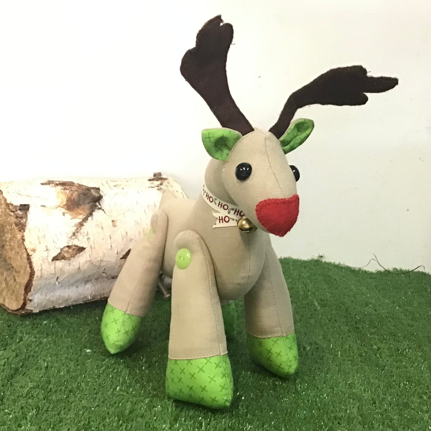 Plush reindeer toy with green feet and red nose on a grassy surface with a log in the background.