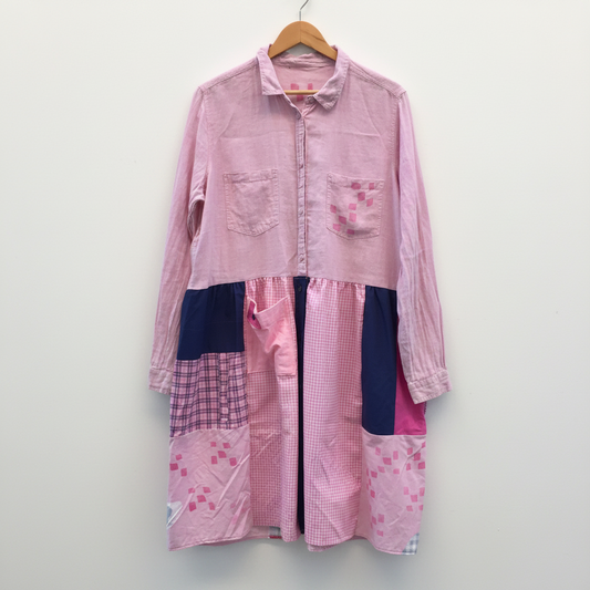 A pink patchwork button-up shirt dress hanging on a wooden hanger against a white painted brick wall. The dress features a light pink corduroy-like upper section, a pink and white gingham patterned midsection, and navy blue and pink patterned patchwork lower panels.