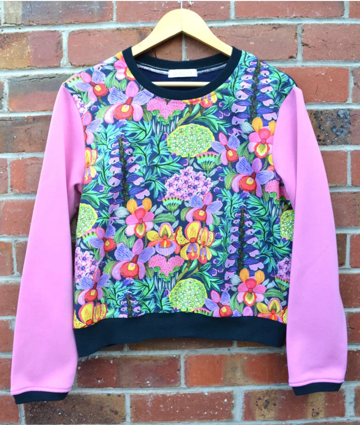 Colorful floral-patterned sweatshirt with pink sleeves on a hanger against a brick wall.