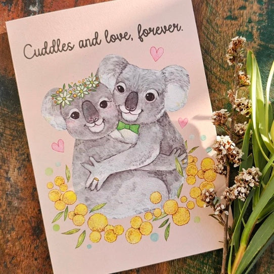 Card with koala illustration and 'Cuddles and love, forever' text on a wooden surface with flowers.