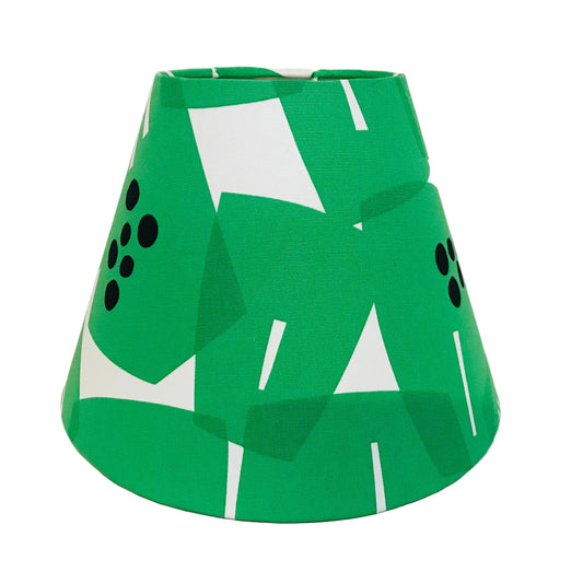 A green fabric lampshade with a bold abstract geometric pattern of white shapes and black dots, photographed against a plain off-white background.