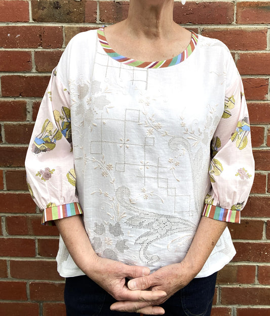 A woman wearing a white embroidered blouse with pastel pink sleeves featuring a floral print and striped trim, standing in front of a red brick wall.