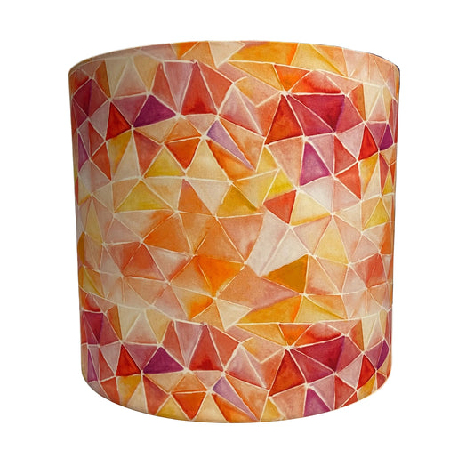 A cylindrical lampshade featuring a colorful watercolor geometric pattern of triangles in orange, yellow, red, and purple on a cream background, displayed on a white table with a workshop or office background.