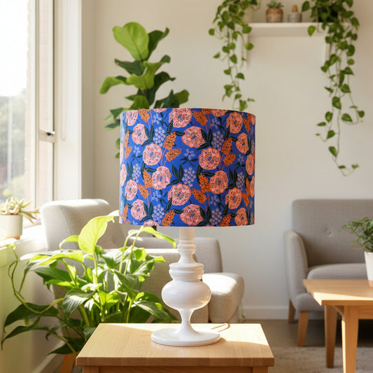 A table lamp with a white pedestal base and a cylindrical drum shade featuring a vibrant floral pattern in blue, pink, and orange, styled on a wooden side table in a bright, modern living room.