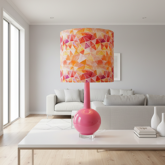 A modern table lamp with a glossy hot pink spherical base and a cylindrical drum shade featuring a colorful geometric triangle pattern in orange, yellow, and pink, styled on a white coffee table in a bright, contemporary living room.