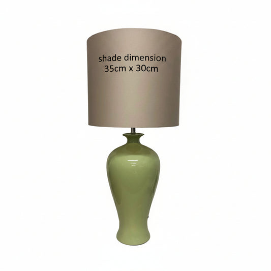 A table lamp with a glossy light green ceramic base and a large taupe-beige fabric drum shade, standing on a white surface against a white wall.