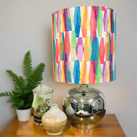 A colorful geometric patterned lampshade on a reflective silver lamp base, placed on a wooden table with decorative items and a potted plant in the background.