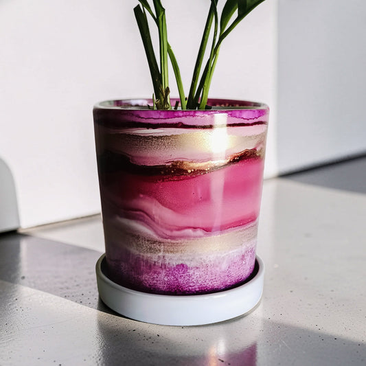 A cylindrical planter with a glossy marbled pink, purple, and beige pattern, holding green plant leaves, sitting on a white saucer on a gray tabletop with strong sunlight and shadows.