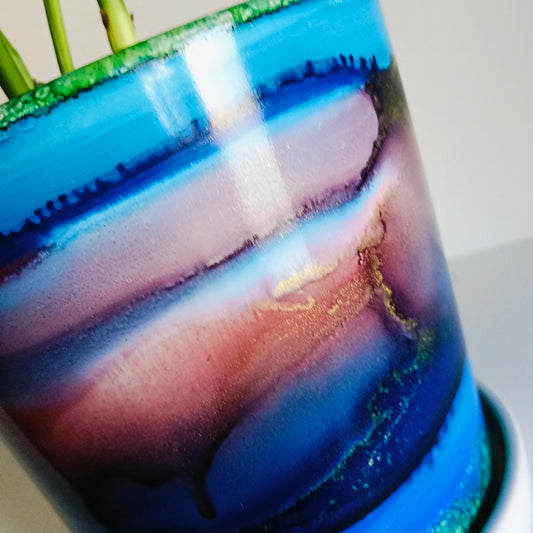 A close-up of a glossy ceramic pot with a marbled abstract design in shades of blue, purple, pink, and gold, with green plant stems visible at the rim.