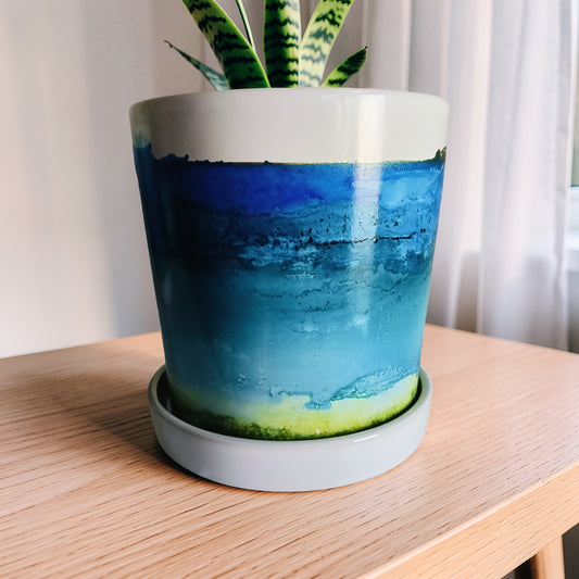 A ceramic planter with a blue and teal watercolor landscape design, sitting on a matching saucer on a light wood table.