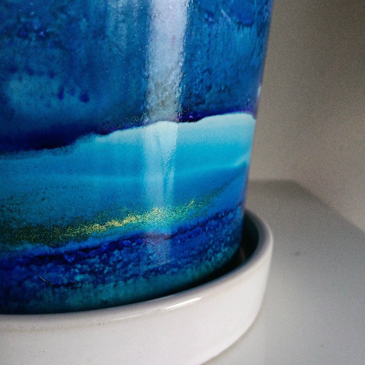 A close-up of a glossy ceramic cup or tumbler with a blue and turquoise landscape-style glaze, resting on a white saucer against a neutral light gray background.