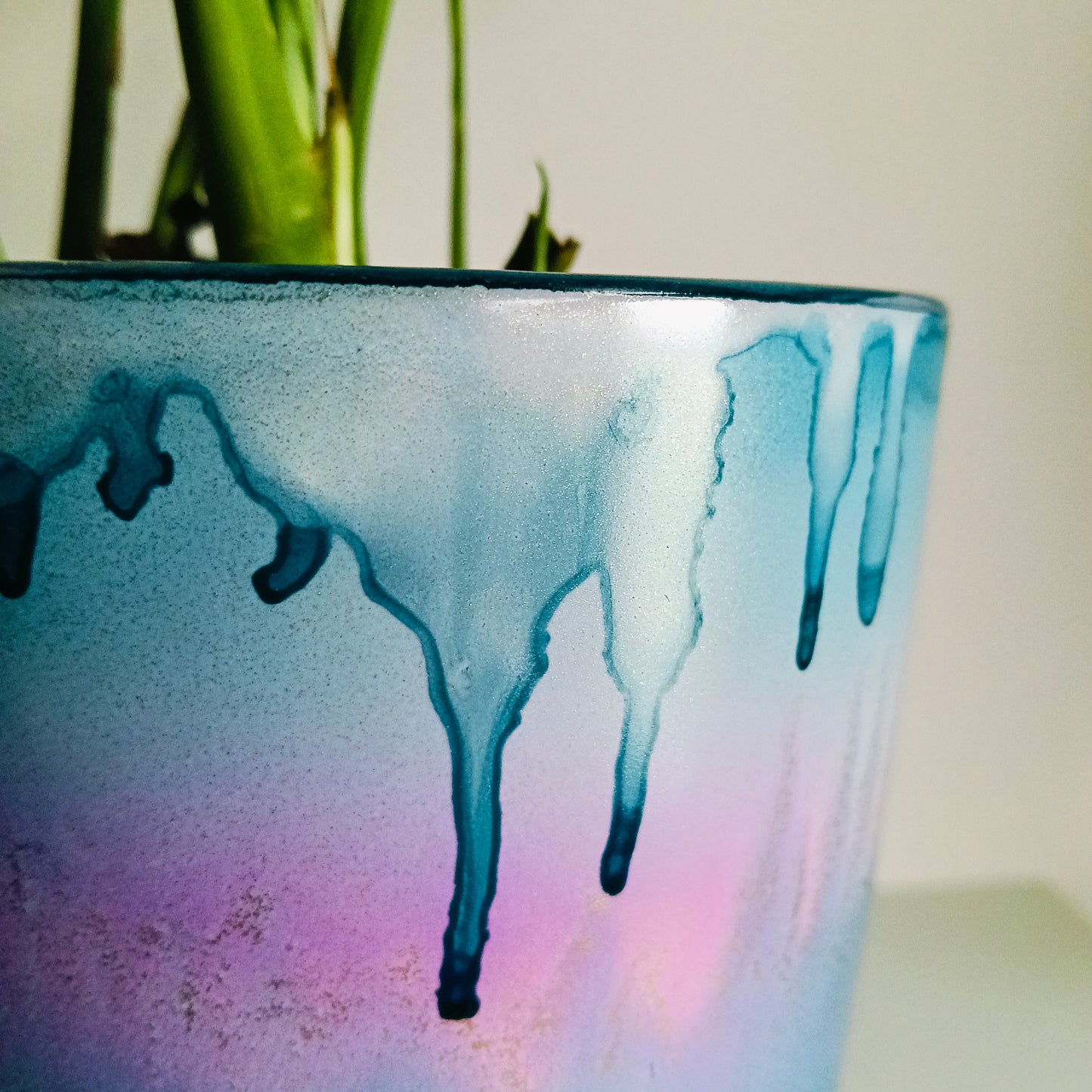 A close-up of a glazed ceramic planter with a blue, purple, and pink drip design, containing green plant stems against a light background.