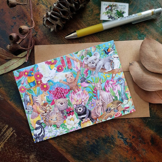 A colorful greeting card featuring Australian wildlife illustrated among flora, laid on a wooden surface with a brown envelope and a pen.