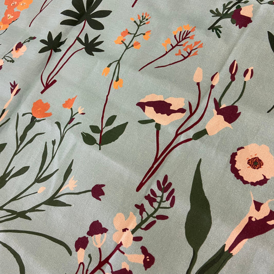 A close-up of a floral fabric print featuring dark green leaves, peach and burgundy flowers, and orange blossoms on a muted sage green background.