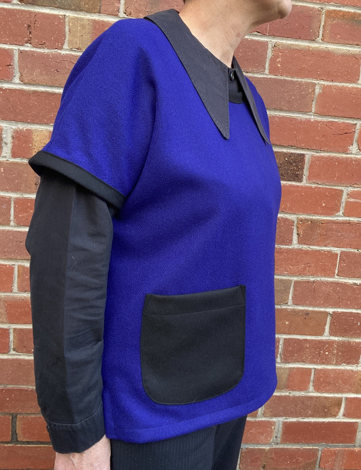 Person wearing a royal blue with blacktrim and pocket wool vest standing against a brick wall