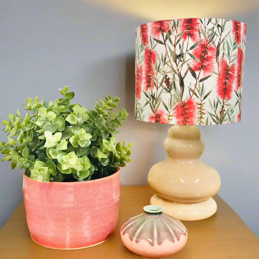 A ceramic table lamp with a pink base and a shade featuring a red and green floral pattern. The lamp is placed next to a potted plant with green leaves.