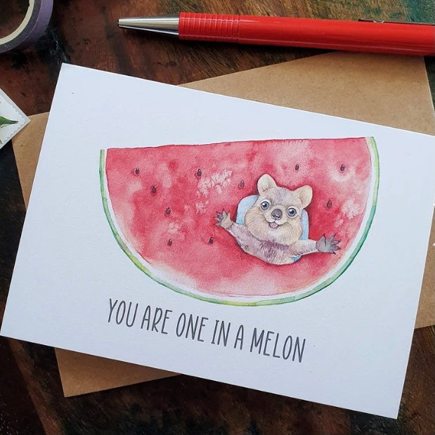 Card with a watermelon slice and bear illustration with text 'You are one in a melon' on a wooden surface.