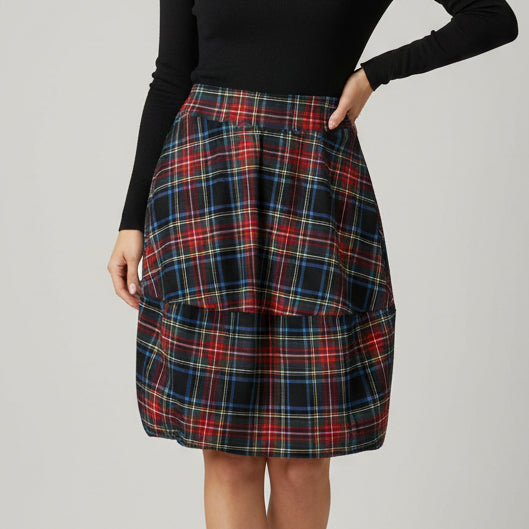 A pleated plaid skirt in a red, black, blue, and yellow tartan pattern, hanging from a wooden hanger against a plain off-white wall.