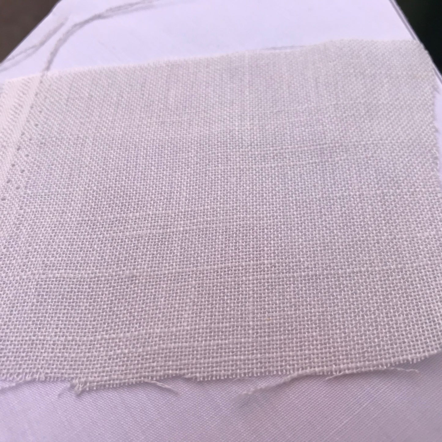 A close-up of a square piece of light beige woven fabric, likely linen or burlap, laid flat on top of a white textured background.