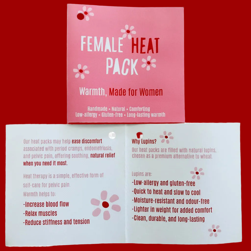 A pink card labeled 'FEMALE HEAT PACK' is placed on top of an unfolded white informational card with red and pink text and flower graphics, both on a wooden surface.