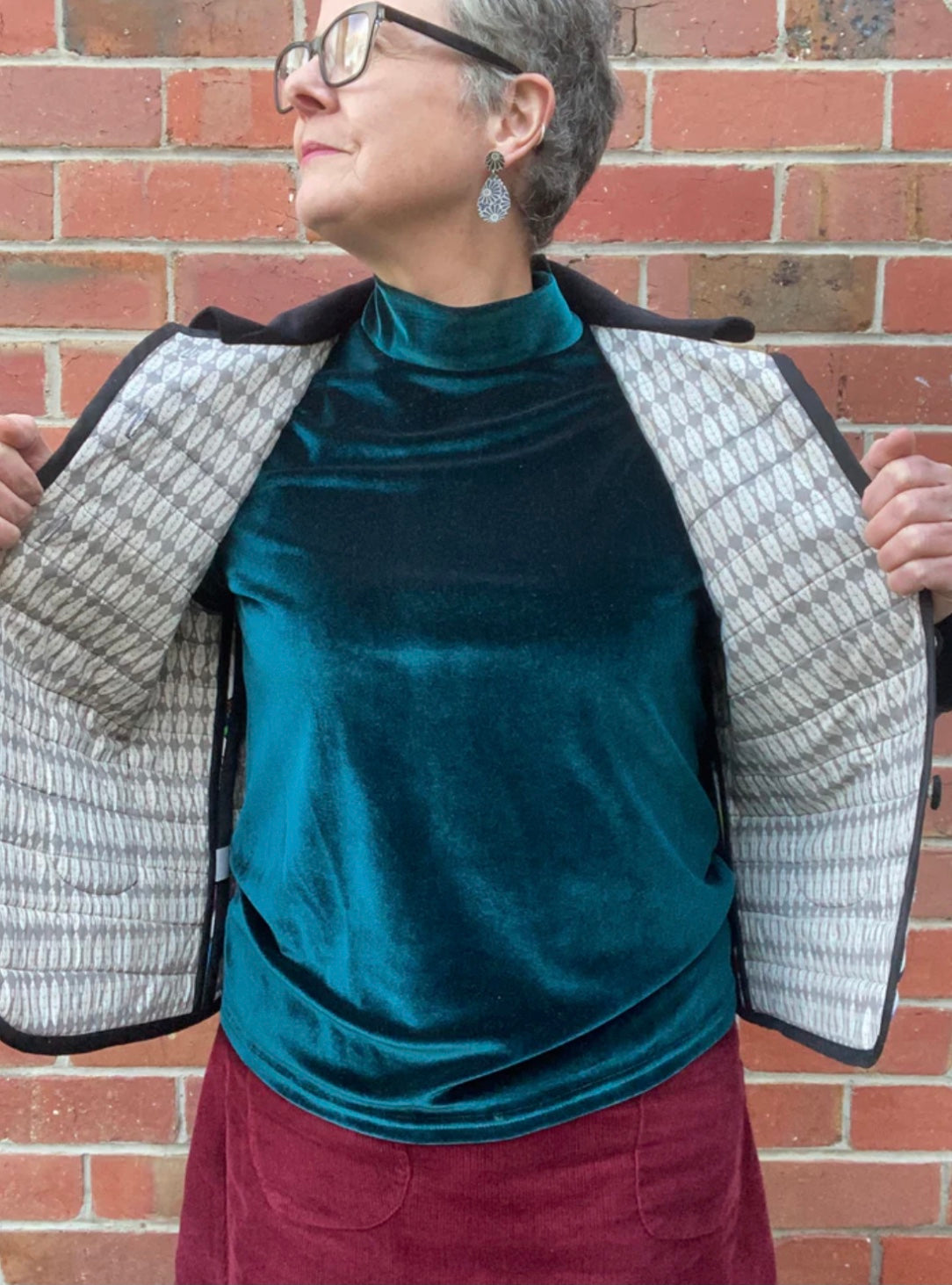 Person wearing a teal velvet top and maroon skirt with a textured gray jacket against a brick wall.