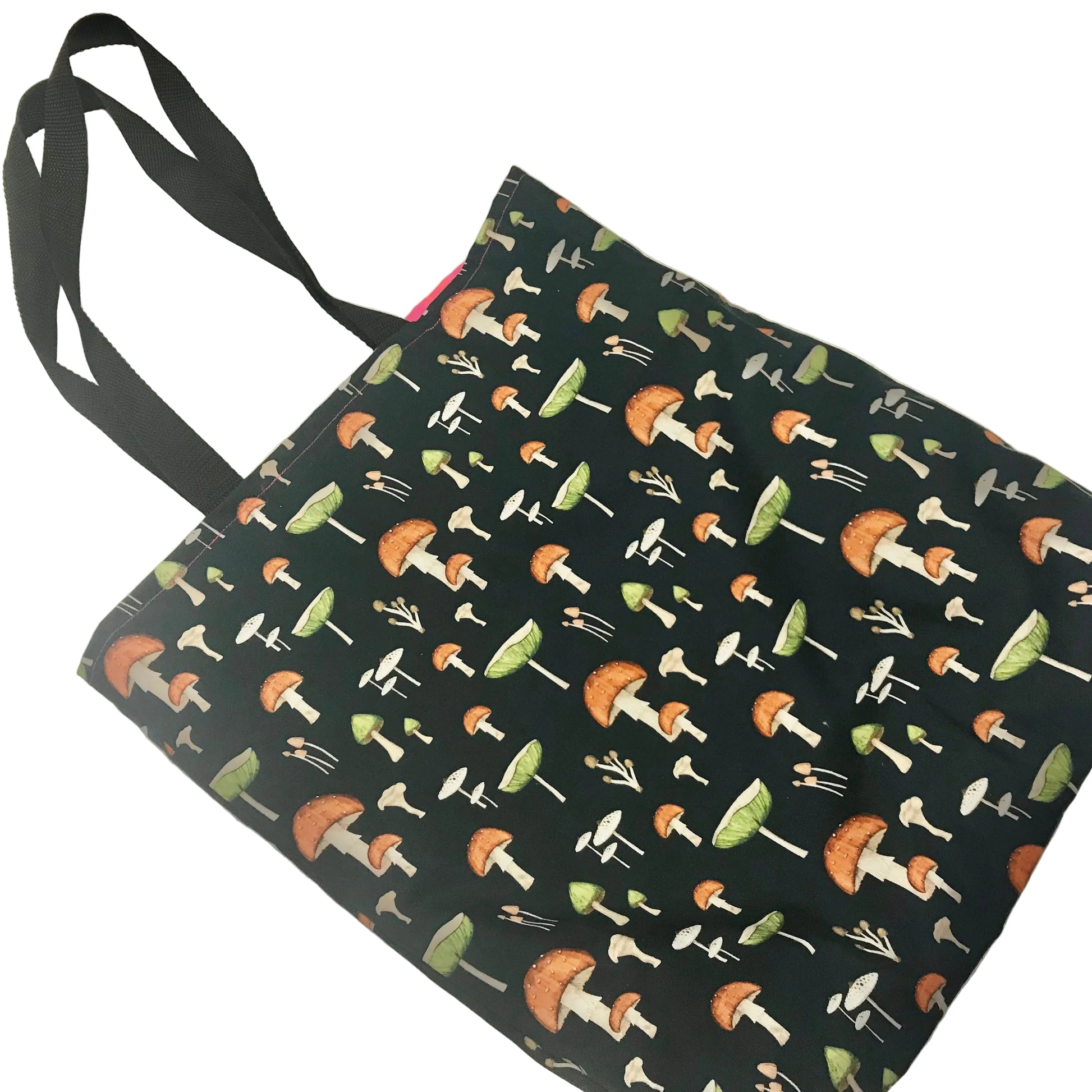 A black fabric tote bag with a repeating mushroom print in orange, green, and white, featuring two black shoulder straps, photographed on a light beige surface.