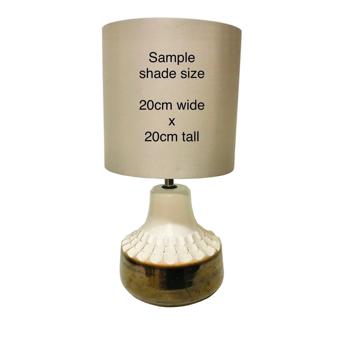 A table lamp with a cylindrical beige shade printed with the text 'Sample shade size 20cm wide x 20cm tall', set on a white surface against a plain off-white wall.