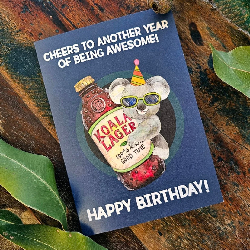 Birthday card with a koala holding a bottle labeled 'Koala Lager' on a wooden surface with leaves.