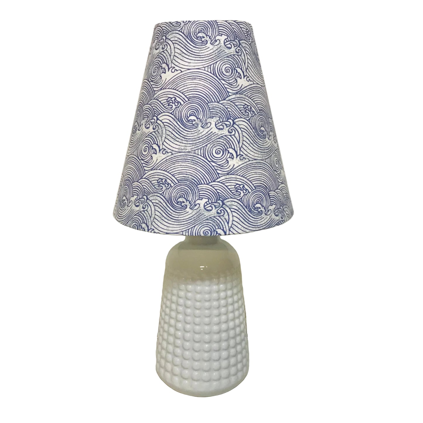 A table lamp with a white textured glass base and a tapered fabric shade featuring a blue and white wave pattern, set against a plain off-white wall.