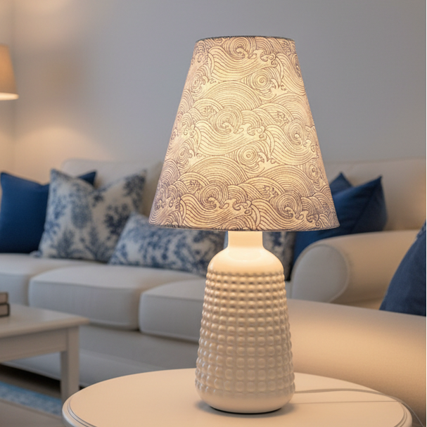 A table lamp with a white textured glass base and a tapered fabric shade featuring a blue and white wave pattern, set against a plain off-white wall.