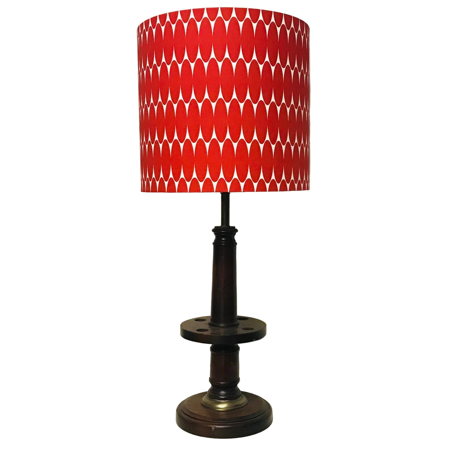 A table lamp with a red and white geometric patterned drum shade and a dark brown wooden base, shown standing on a white surface against an off-white wall.
