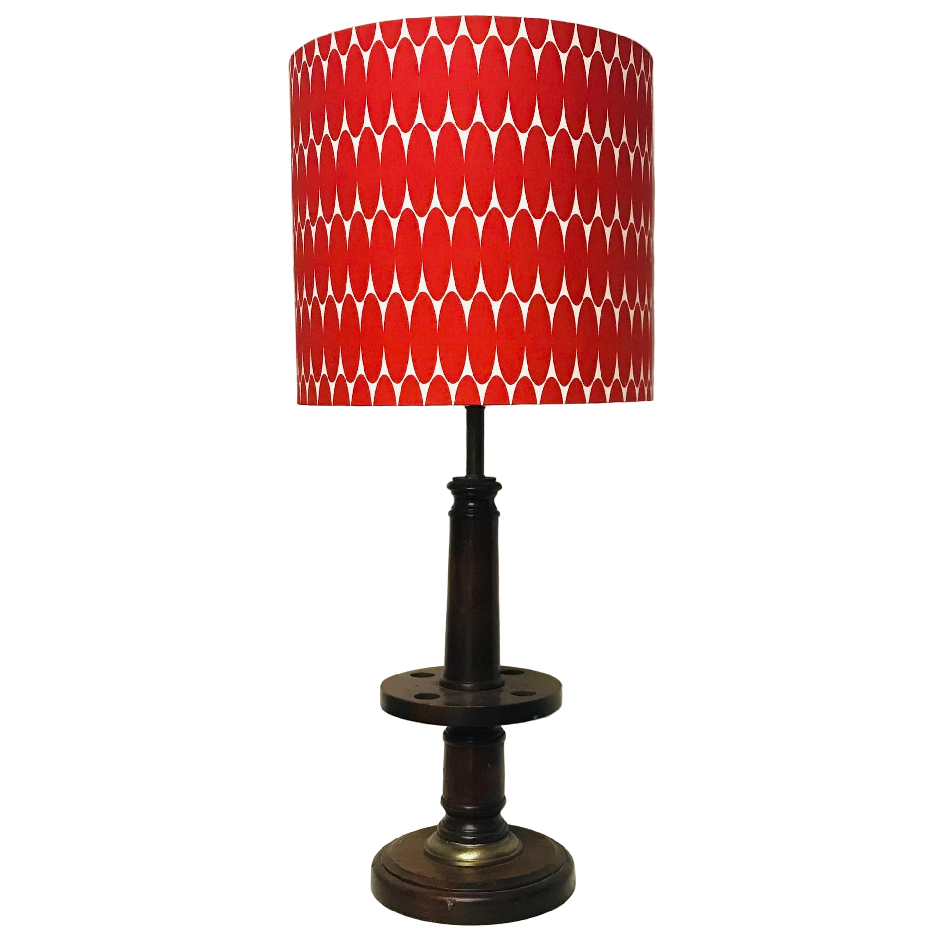 A table lamp with a red and white geometric patterned drum shade and a dark brown wooden base, shown standing on a white surface against an off-white wall.