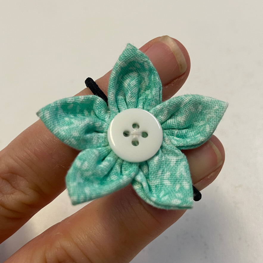 Green fabric hairtie flower with a button center held between fingers on a white background
