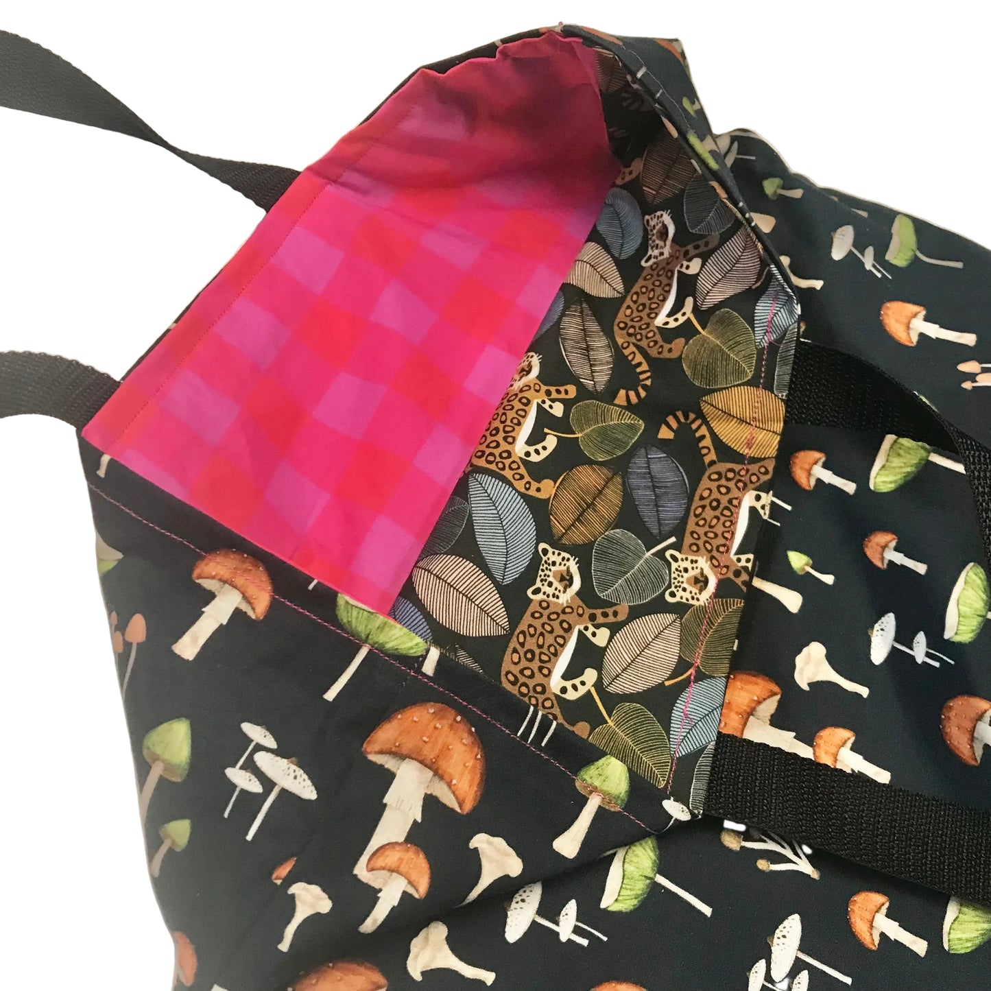 Close-up of a fabric tote bag showing multiple printed panels: a black background with white, orange, and green mushrooms, a leopard-and-leaf print panel, and a bright pink plaid panel. Black webbing straps are visible.